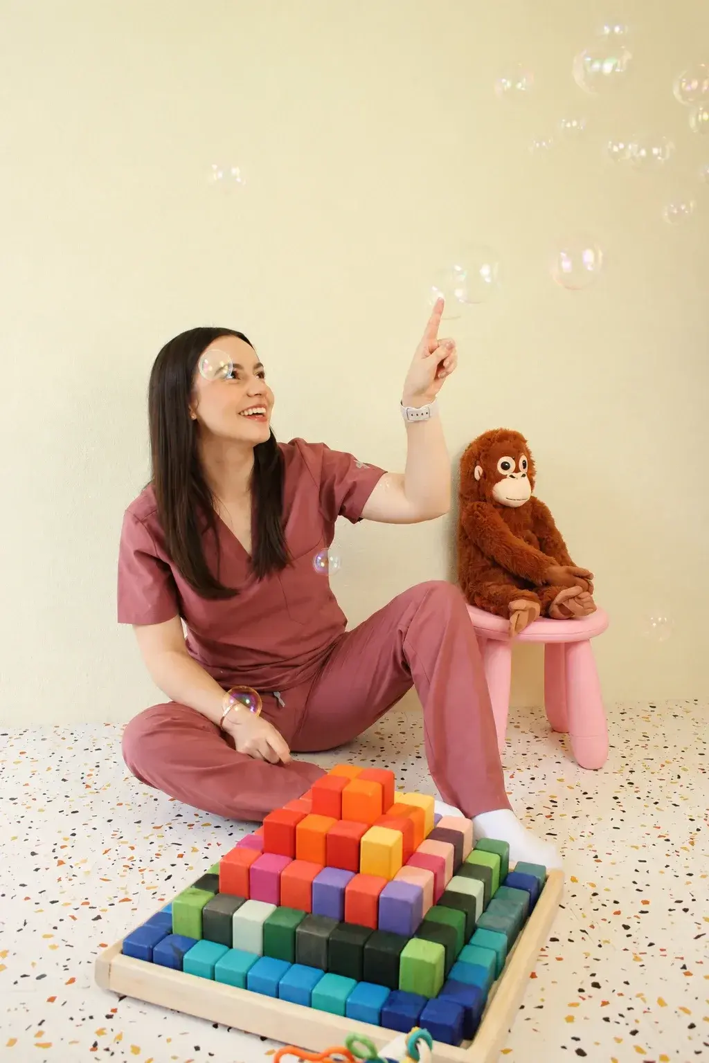 Dani sentada en el piso señalando burbujas junto a una pirámide de bloques de colores y un peluche