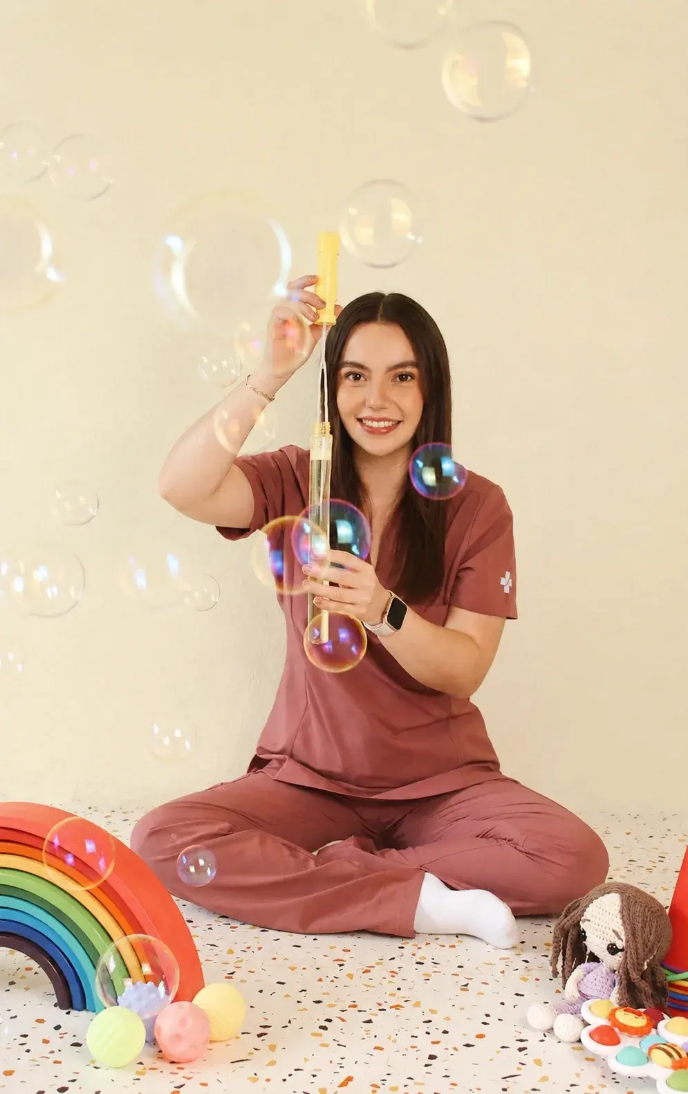 Dani sentada en el piso haciendo burbujas, rodeada de un arcoíris de madera, pelotas sensoriales y una muñeca tejida